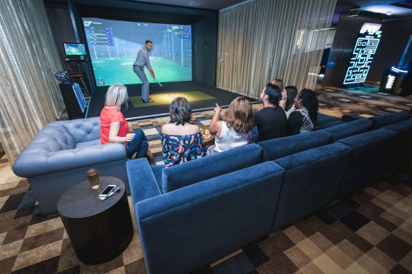 A group of people sits on couches watching a golf simulation on a large screen in a modern, dimly lit entertainment room.