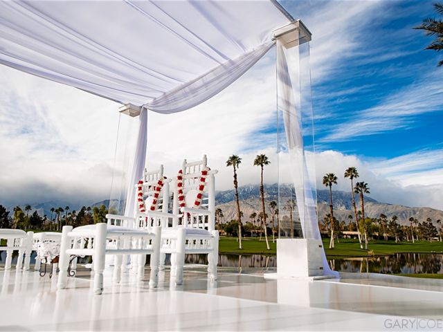 White outdoor wedding setup with chairs, decorated canopy, palm trees, mountains, and clouds in the background.