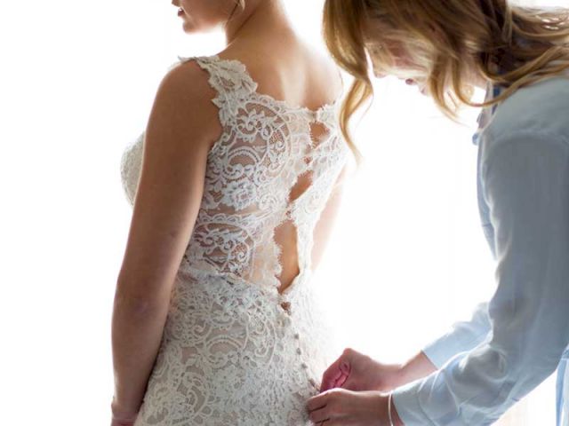 A bride in a lace dress is getting ready as another person helps button up the back.