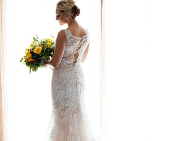 A bride in a lace wedding dress stands by a bright window, holding a bouquet with yellow flowers, looking thoughtfully at it.