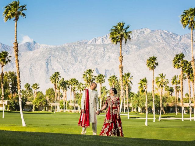 A couple in traditional attire stands on a lush landscape with palm trees, mountains, and a blue sky in the background.