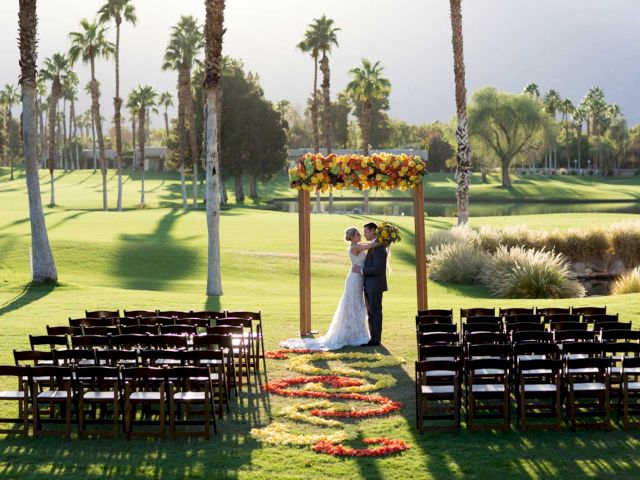 A couple stands under a flower-adorned archway on a grassy outdoor aisle, surrounded by empty chairs, with a scenic backdrop of palm trees.