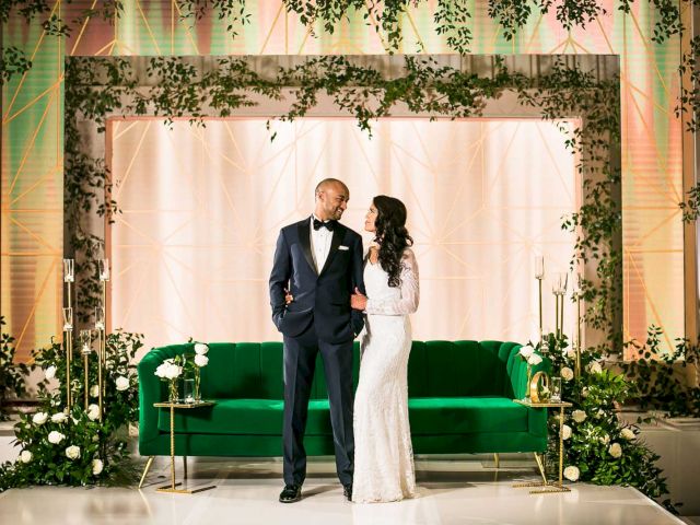 A couple in formal attire is standing in a decorative setting with a green sofa and floral arrangements.