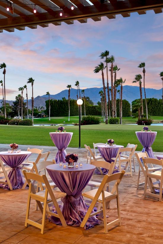 Outdoor event setup with tables draped in purple, chairs, and lights under a pergola, with a scenic backdrop of palm trees and mountains.