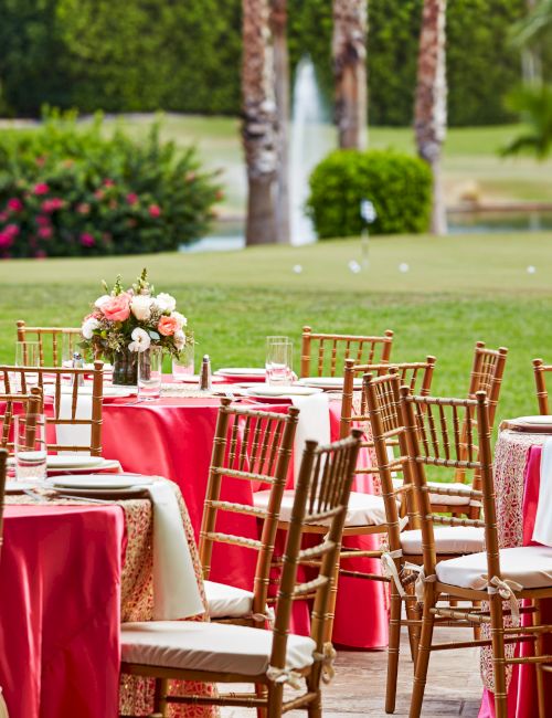 Outdoor event setup with tables covered in pink cloths, floral centerpieces, and gold chairs on a green lawn by a fountain.