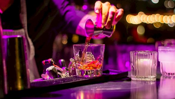 A bartender is pouring a drink into a glass with ice, surrounded by bar tools, under vibrant purple lighting.