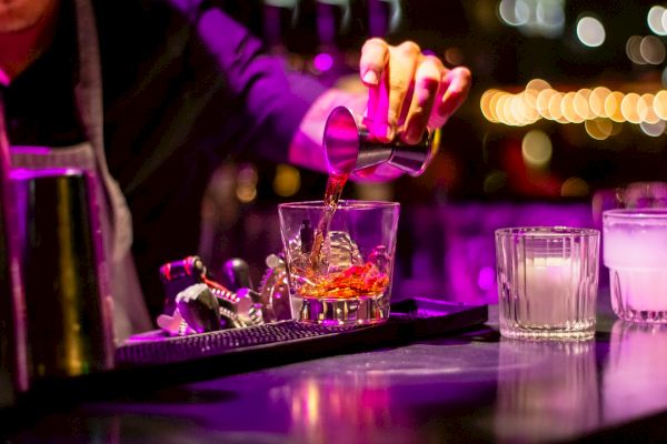 A bartender is pouring a drink into a glass with ice, surrounded by bar tools, under vibrant purple lighting.