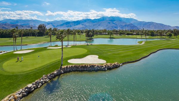 A scenic golf course with lush greens, sand traps, palm trees, and a lake, set against a backdrop of distant mountains under a clear blue sky.