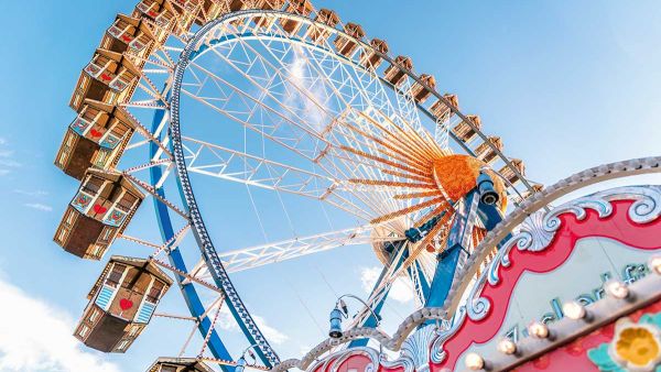 The image shows a colorful Ferris wheel against a blue sky, with ornate details at the base, likely part of a fairground or amusement park.