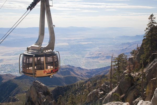 A rotating aerial tramway cable car travels over rugged mountain terrain with a scenic valley view below in the background.