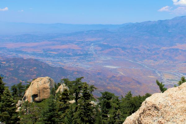 Rocky ledge overlooks forested landscape and distant mountains with a clear blue sky above.