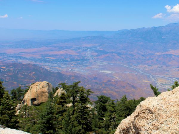 Rocky ledge overlooks forested landscape and distant mountains with a clear blue sky above.