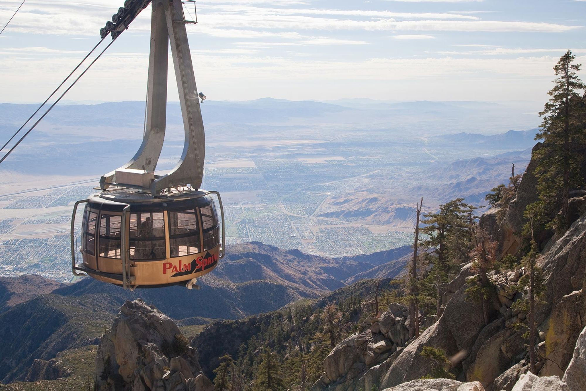 A cable car ascends over rocky terrain with expansive views of a valley and mountains under a clear sky.