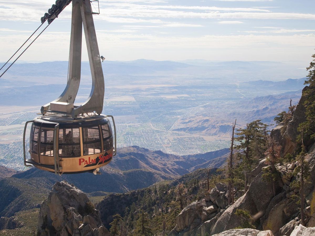 A cable car ascends over a rocky mountain landscape with expansive views of a valley and distant mountains.