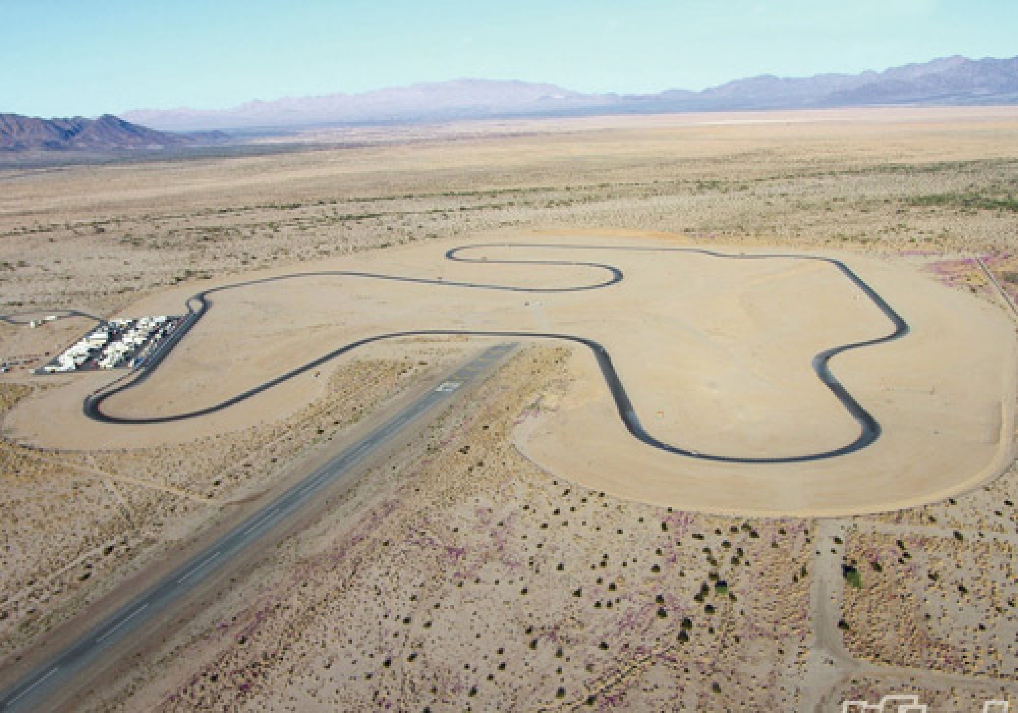 The image shows a desert landscape with a winding asphalt track, possibly a test track or raceway, surrounded by open, barren terrain.