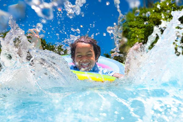 A child joyfully splashes in a pool, floating on an inflatable ring under a clear blue sky with water droplets in motion.