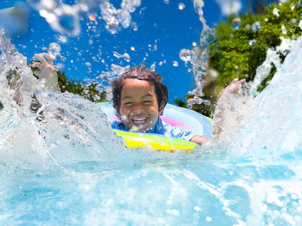 A child joyfully splashes in a pool, floating on an inflatable ring under a clear blue sky with water droplets in motion.