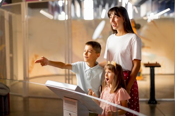 A family is enjoying an interactive exhibit, with a woman and two kids looking excitedly and pointing inside a modern setting.