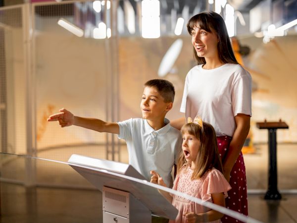 A family is enjoying an interactive exhibit, with a woman and two kids looking excitedly and pointing inside a modern setting.