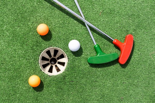 The image shows two mini golf putters, orange and white golf balls, and a hole on artificial green turf.
