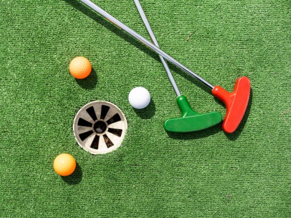 The image shows two mini golf putters, orange and white golf balls, and a hole on artificial green turf.