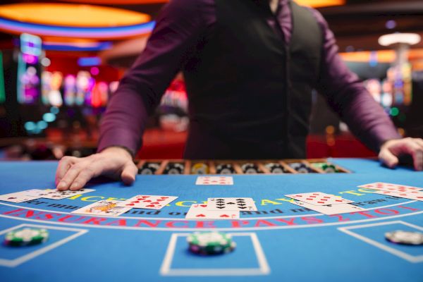 A casino dealer stands at a blackjack table with several cards and chips. The background is filled with glowing slot machines.