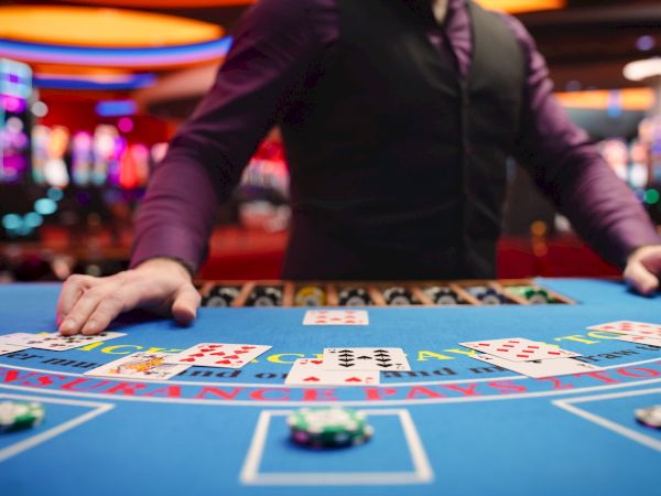 A casino dealer stands at a blackjack table with several cards and chips. The background is filled with glowing slot machines.