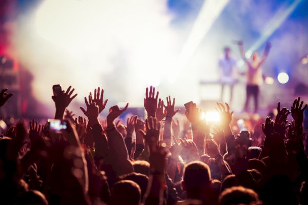 A crowd with raised hands at a concert, colorful lights illuminate the background as performers are on stage.