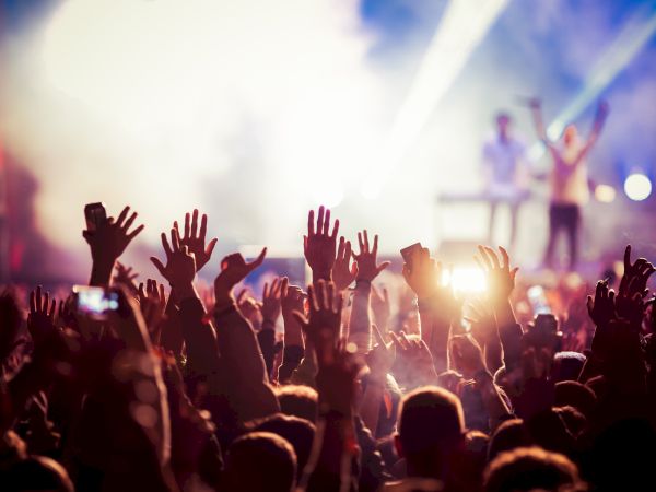 A crowd with raised hands at a concert, colorful lights illuminate the background as performers are on stage.