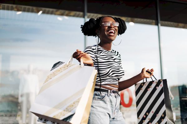 A person smiling and holding shopping bags, wearing a striped shirt and sunglasses, stands in front of a store window.