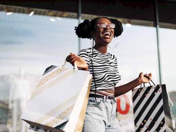 A person smiling and holding shopping bags, wearing a striped shirt and sunglasses, stands in front of a store window.