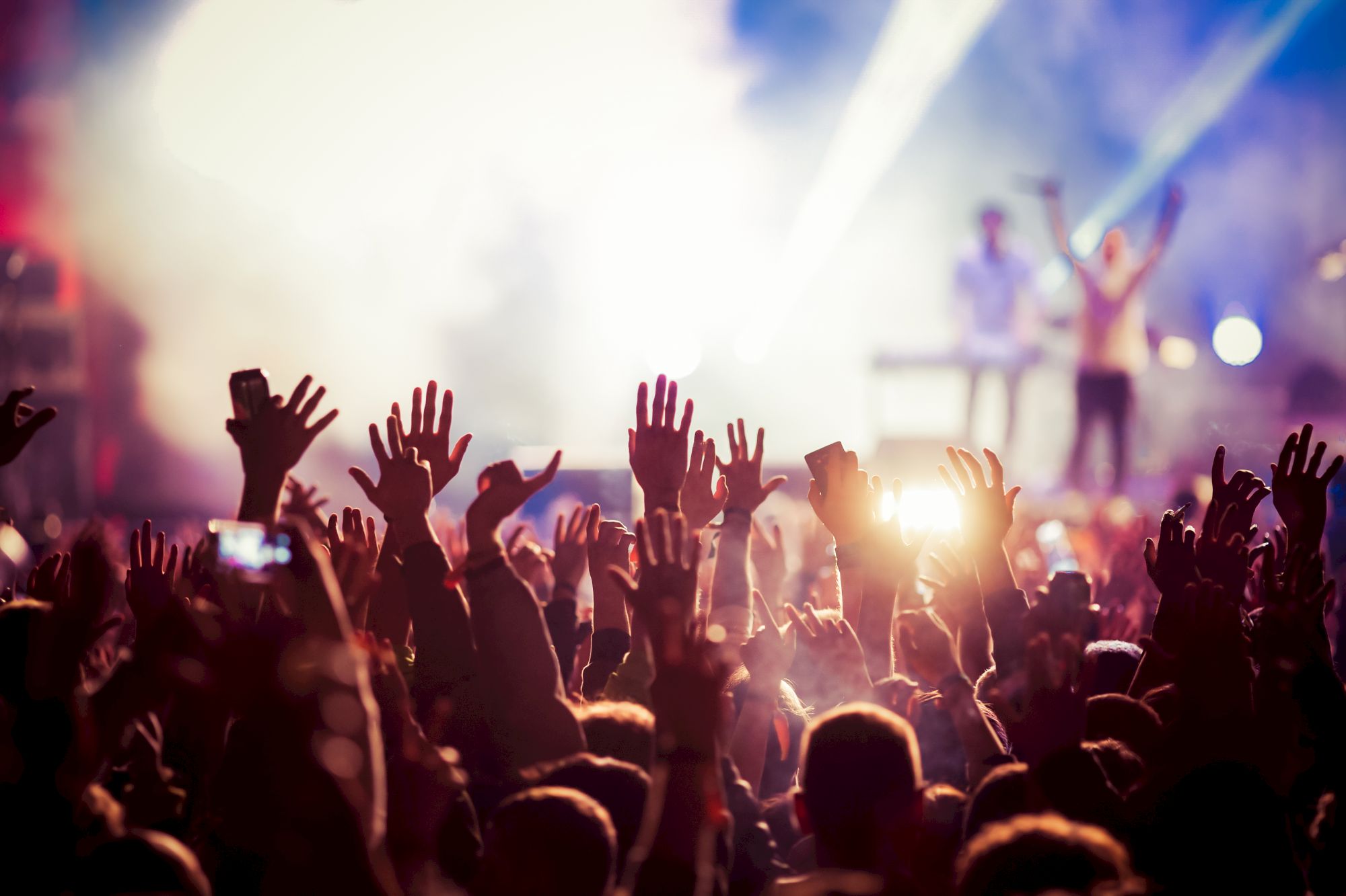 A crowd at a concert with hands raised, with bright stage lights and a performer in the background.