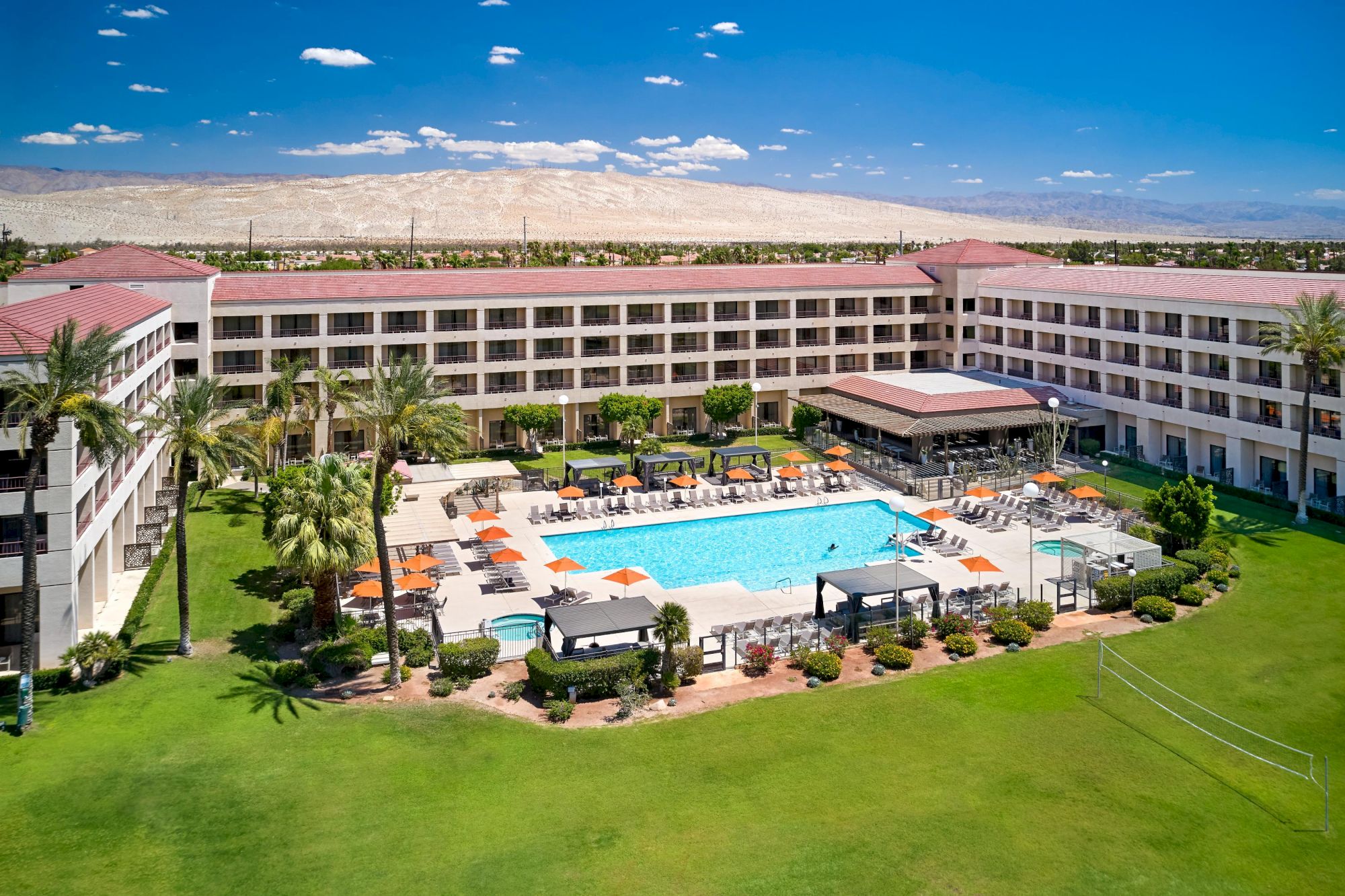This image shows a hotel with a large central pool, surrounded by lounge chairs, palm trees, and a well-maintained lawn.