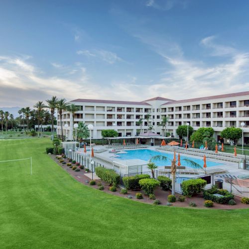 A hotel with a large pool, surrounded by greenery and palm trees, under a clear sky.