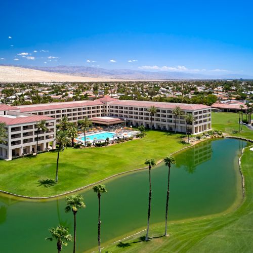 A resort with a pool, surrounded by green lawns, palm trees, a golf course, and a body of water; mountains visible in the background.