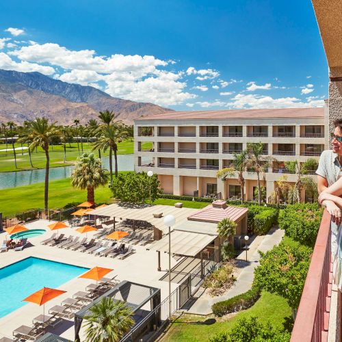 A couple enjoys a scenic view from a balcony overlooking a hotel pool, golf course, and mountains under a sunny sky.