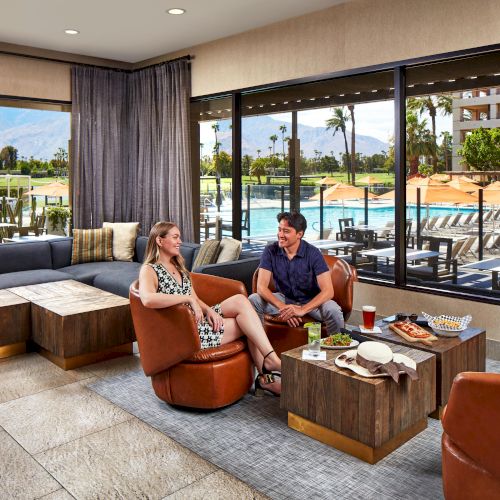 A couple sits in a cozy lounge with large windows showing a poolside view and mountains in the background.