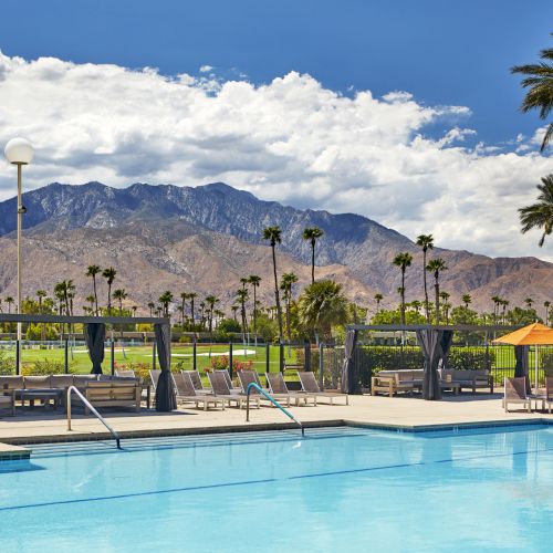 A pool with lounge chairs and cabanas, set against a backdrop of mountains and palm trees, under a partly cloudy sky.