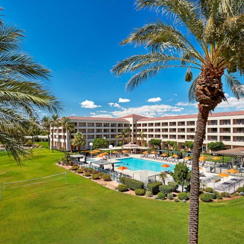 A resort with a large pool, surrounded by palm trees and green lawns, featuring orange umbrellas and a building in the background.