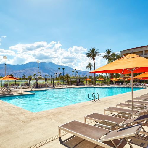 A sunny pool area with loungers and orange umbrellas, surrounded by palm trees and mountains in the distance.
