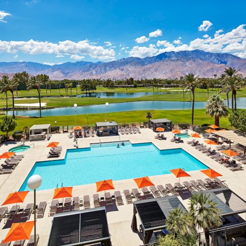 A resort-style swimming pool surrounded by lounge chairs and orange umbrellas, with a golf course and mountains in the background.