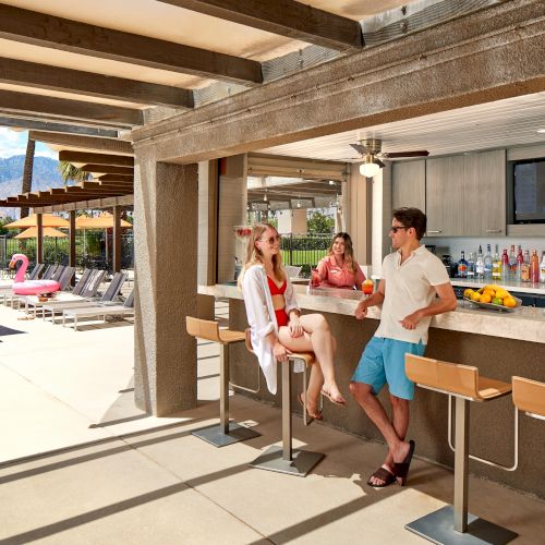 Outdoor poolside bar with people enjoying drinks, surrounded by sun loungers and umbrellas in a sunny setting.