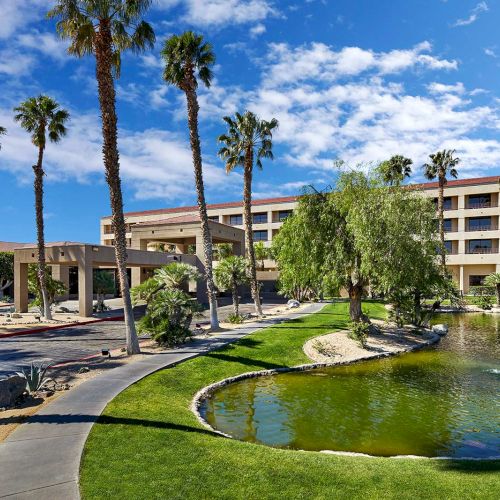 A resort-style building surrounded by palm trees, a pond with fountains, and a well-kept lawn under a blue sky with clouds.