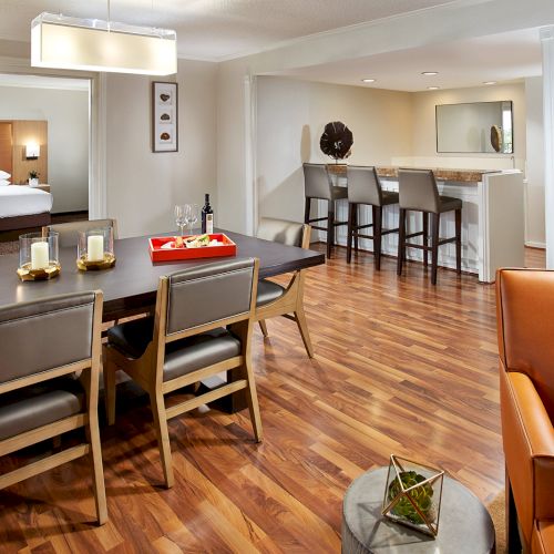 A modern hotel suite with a dining table, bar stools, bedroom, and a TV. Warm lighting and wooden flooring complete the cozy setting.