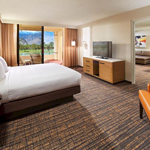 A spacious hotel room with a bed, TV, armchair, and a view of mountains through a sliding door, leading to a balcony.