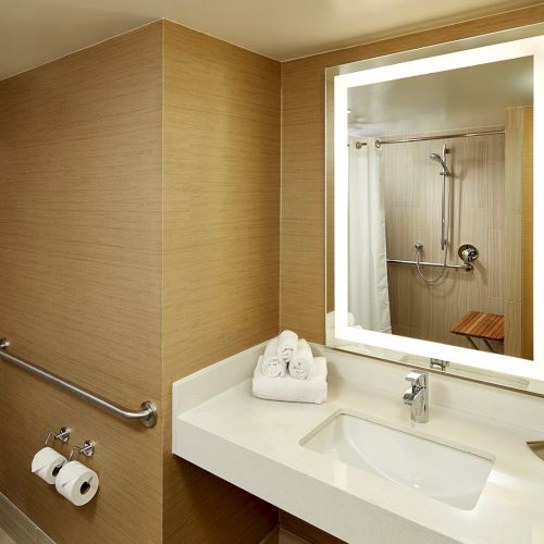 A modern bathroom with a lit mirror, sink, toiletries, and shower in the reflection. There's a toilet and decorative wallpaper.