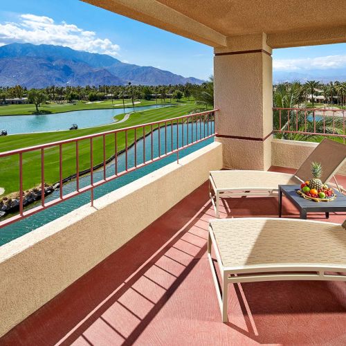 A sunny balcony with lounge chairs and fruit overlooks a golf course with a lake and mountains in the background.