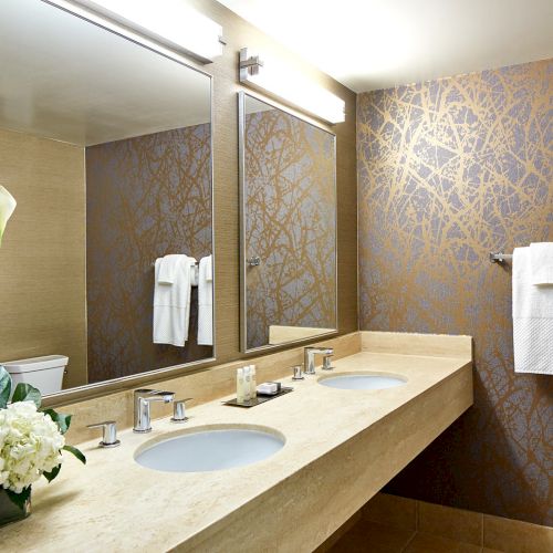Modern bathroom with dual sinks, wall mirror, geometric wallpaper, white towels, and a floral arrangement in a vase on a marble countertop.