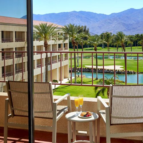 A balcony with two chairs, a table with drinks and food, overlooking a golf course, palm trees, a lake, and mountains in the distance.