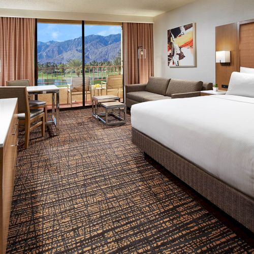 A hotel room with a bed, TV, sofa, and table by a window showing mountains. The decor is modern and neutral in tone.